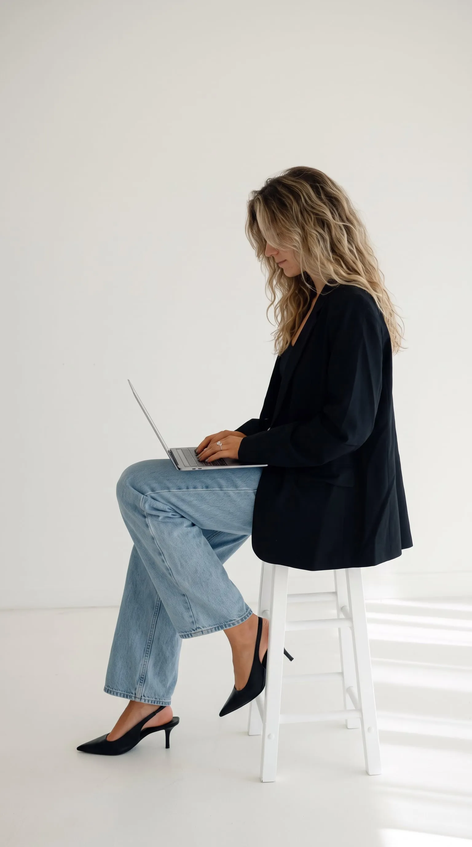 A woman working on a laptop, sitting on a white stool